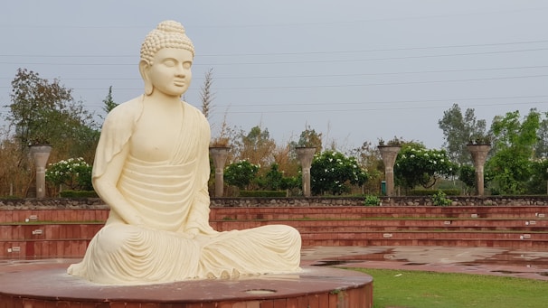 A large, serene statue of Buddha made from a light-colored stone sits on a circular platform in an open area. The statue is surrounded by greenery and trees, with an amphitheater-style seating structure made from red stone in the background. The environment is peaceful, with overcast skies above.
