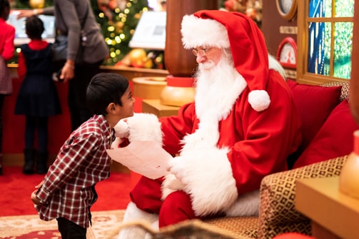 A child in a red and white plaid shirt is interacting enthusiastically with a person dressed as Santa Claus, who is sitting on a cozy, decorated chair. The Santa figure is holding a letter, presumably a wish list, and is smiling at the child. The background is festively decorated with a Christmas tree and other ornaments, suggesting a holiday setting.