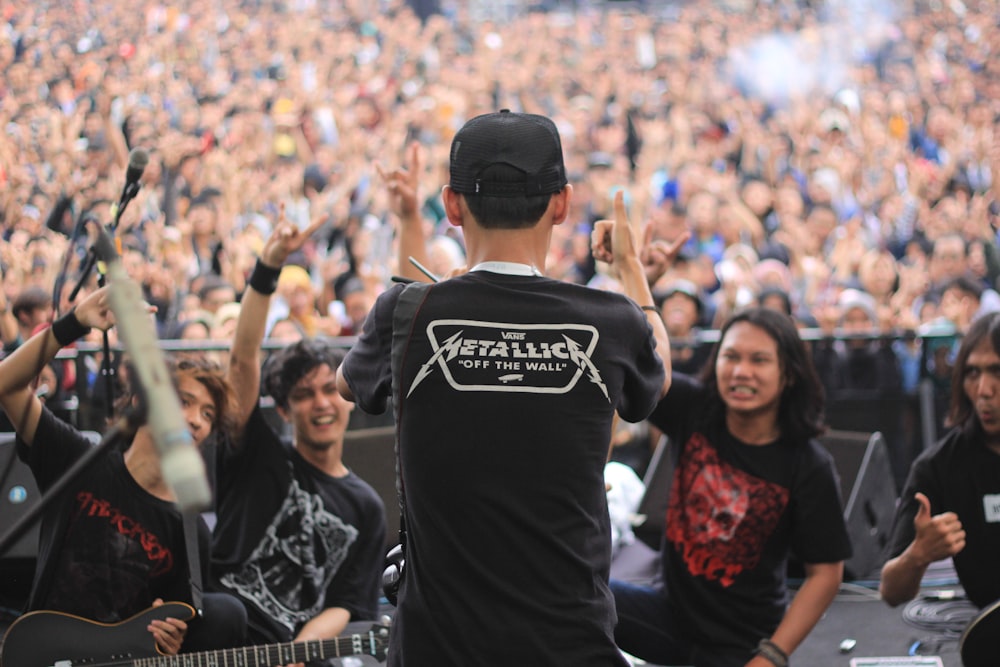 man wearing black and white Metallica t-shirt doing thumbs up hand gesture in front of band performer on top of stage
