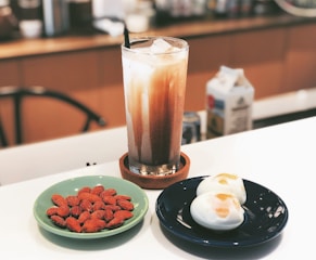 A tall glass filled with iced coffee or milk tea sits atop a coaster. Next to it, on a light green plate, are several almonds. On a dark blue plate, there are two halved boiled eggs with runny yolks. A milk carton can be seen in the blurred background.