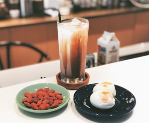 A tall glass filled with iced coffee or milk tea sits atop a coaster. Next to it, on a light green plate, are several almonds. On a dark blue plate, there are two halved boiled eggs with runny yolks. A milk carton can be seen in the blurred background.