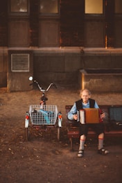 An elderly person with long hair playing an accordion sits on a bench next to a bicycle with an attached basket. The setting is an urban outdoor environment with a brick wall and windows in the background. Soft evening light casts shadows and creates a warm, nostalgic atmosphere.