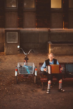 An elderly person with long hair playing an accordion sits on a bench next to a bicycle with an attached basket. The setting is an urban outdoor environment with a brick wall and windows in the background. Soft evening light casts shadows and creates a warm, nostalgic atmosphere.