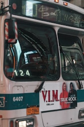 A city bus displays a digital route number and advertisement on its side. The bus is cream and teal with a prominent V.I.M. clothing advertisement featuring two models. The front section of the bus shows reflections and part of the driver's seat.