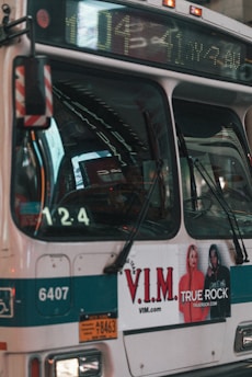 A city bus displays a digital route number and advertisement on its side. The bus is cream and teal with a prominent V.I.M. clothing advertisement featuring two models. The front section of the bus shows reflections and part of the driver's seat.