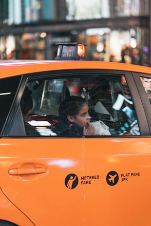 A young person sits inside an orange taxi, looking out the window. The view includes the side of the taxi with metered fare and flat fare JFK markings. The background shows a blurred cityscape with lights and indications of movement.