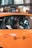 A young person sits inside an orange taxi, looking out the window. The view includes the side of the taxi with metered fare and flat fare JFK markings. The background shows a blurred cityscape with lights and indications of movement.