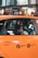A young person sits inside an orange taxi, looking out the window. The view includes the side of the taxi with metered fare and flat fare JFK markings. The background shows a blurred cityscape with lights and indications of movement.