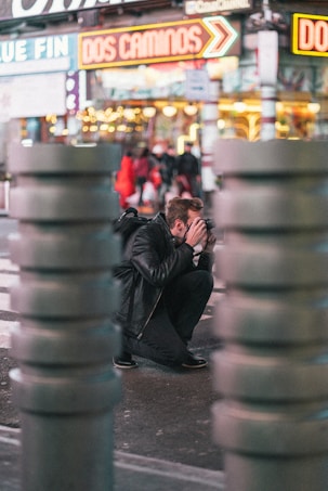 A person crouches on the sidewalk, focused on capturing a photograph with a camera in hand. Neon signs in the background create a vibrant city atmosphere, while blurred foreground objects give a sense of depth.