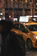 A yellow taxi is parked on a dimly lit city street, with bright advertisements on its rooftop. The background shows a bustling urban environment with neon signs and a blurred pedestrian passing by.