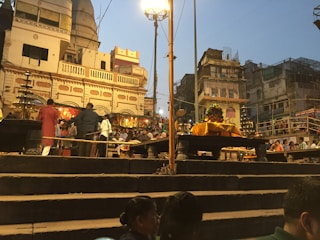 A busy scene at a ghat with people gathered for a religious ceremony. There are steps leading up to a raised platform with offerings and decorations. The buildings in the background have traditional architecture, and the atmosphere is lively with many individuals engaged in various activities. Street lamps illuminate the area as dusk approaches.
