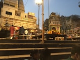 A busy scene at a ghat with people gathered for a religious ceremony. There are steps leading up to a raised platform with offerings and decorations. The buildings in the background have traditional architecture, and the atmosphere is lively with many individuals engaged in various activities. Street lamps illuminate the area as dusk approaches.