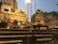 A busy scene at a ghat with people gathered for a religious ceremony. There are steps leading up to a raised platform with offerings and decorations. The buildings in the background have traditional architecture, and the atmosphere is lively with many individuals engaged in various activities. Street lamps illuminate the area as dusk approaches.