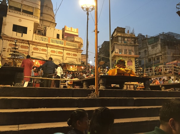 A busy scene at a ghat with people gathered for a religious ceremony. There are steps leading up to a raised platform with offerings and decorations. The buildings in the background have traditional architecture, and the atmosphere is lively with many individuals engaged in various activities. Street lamps illuminate the area as dusk approaches.