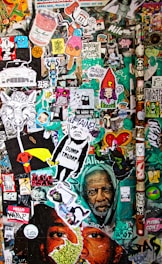 A vibrant collage of street art and stickers covers a wall, featuring various illustrations and messages, including political statements, cartoon characters, and abstract art. The background is filled with layers of overlapping images and graffiti in bright colors, creating a chaotic and lively scene.