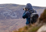 A person with a blue jacket and hood is holding a camera and taking photos in a mountainous area. They have a black backpack and are surrounded by natural landscape with earthy tones.