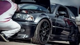A person is kneeling next to a black sports car with the hood open, examining the engine. The car has a sleek design with a glossy finish, featuring sporty rims and a decal on the windshield. The individual is wearing white pants and a patterned shirt with red and white colors.