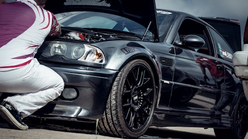 A person is kneeling next to a black sports car with the hood open, examining the engine. The car has a sleek design with a glossy finish, featuring sporty rims and a decal on the windshield. The individual is wearing white pants and a patterned shirt with red and white colors.