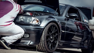 A person is kneeling next to a black sports car with the hood open, examining the engine. The car has a sleek design with a glossy finish, featuring sporty rims and a decal on the windshield. The individual is wearing white pants and a patterned shirt with red and white colors.
