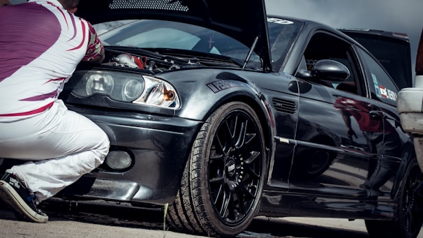 A person is kneeling next to a black sports car with the hood open, examining the engine. The car has a sleek design with a glossy finish, featuring sporty rims and a decal on the windshield. The individual is wearing white pants and a patterned shirt with red and white colors.