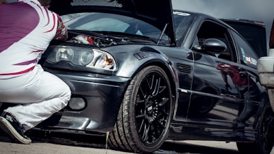 A person is kneeling next to a black sports car with the hood open, examining the engine. The car has a sleek design with a glossy finish, featuring sporty rims and a decal on the windshield. The individual is wearing white pants and a patterned shirt with red and white colors.