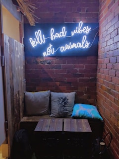 A cozy corner featuring a dark brick wall with a neon sign displaying the phrase 'kill bad vibes not animals' in white cursive letters. Below the sign is a wooden bench adorned with three cushions: one beige, one with a pineapple design, and one blue with a white cloud-like pattern. The setting has a rustic and warm ambiance.
