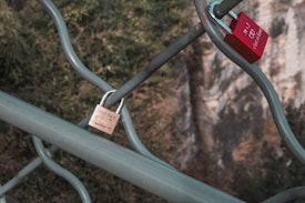 Two padlocks are attached to a metal fence overlooking a blurred background of greenery and rocky terrain. One lock is red with a white engraving, and the other is a light tan color with writing indicating themes of friendship.