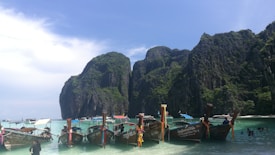 Several traditional long-tail boats are lined up near a scenic beach. The clear turquoise water and the lush, towering cliffs in the background create a picturesque tropical setting. A few people are wading in the shallow water.