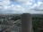 A tall, partially constructed high-rise building stands prominently against a backdrop of a sprawling urban landscape. The foreground shows the building surrounded by other mid-rise structures and various residential areas. The sky is filled with scattered clouds, offering a mix of sunlight and shadow across the scene.