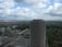 A tall, partially constructed high-rise building stands prominently against a backdrop of a sprawling urban landscape. The foreground shows the building surrounded by other mid-rise structures and various residential areas. The sky is filled with scattered clouds, offering a mix of sunlight and shadow across the scene.