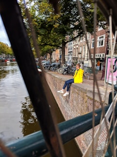 A person in a bright yellow jacket sits on the edge of a brick canal wall, with their legs dangling over the water. Bicycles are lined up along the path next to a row of typical Dutch-style buildings. The scene is framed by blurry bicycle spokes and green foliage from trees appears in the background.