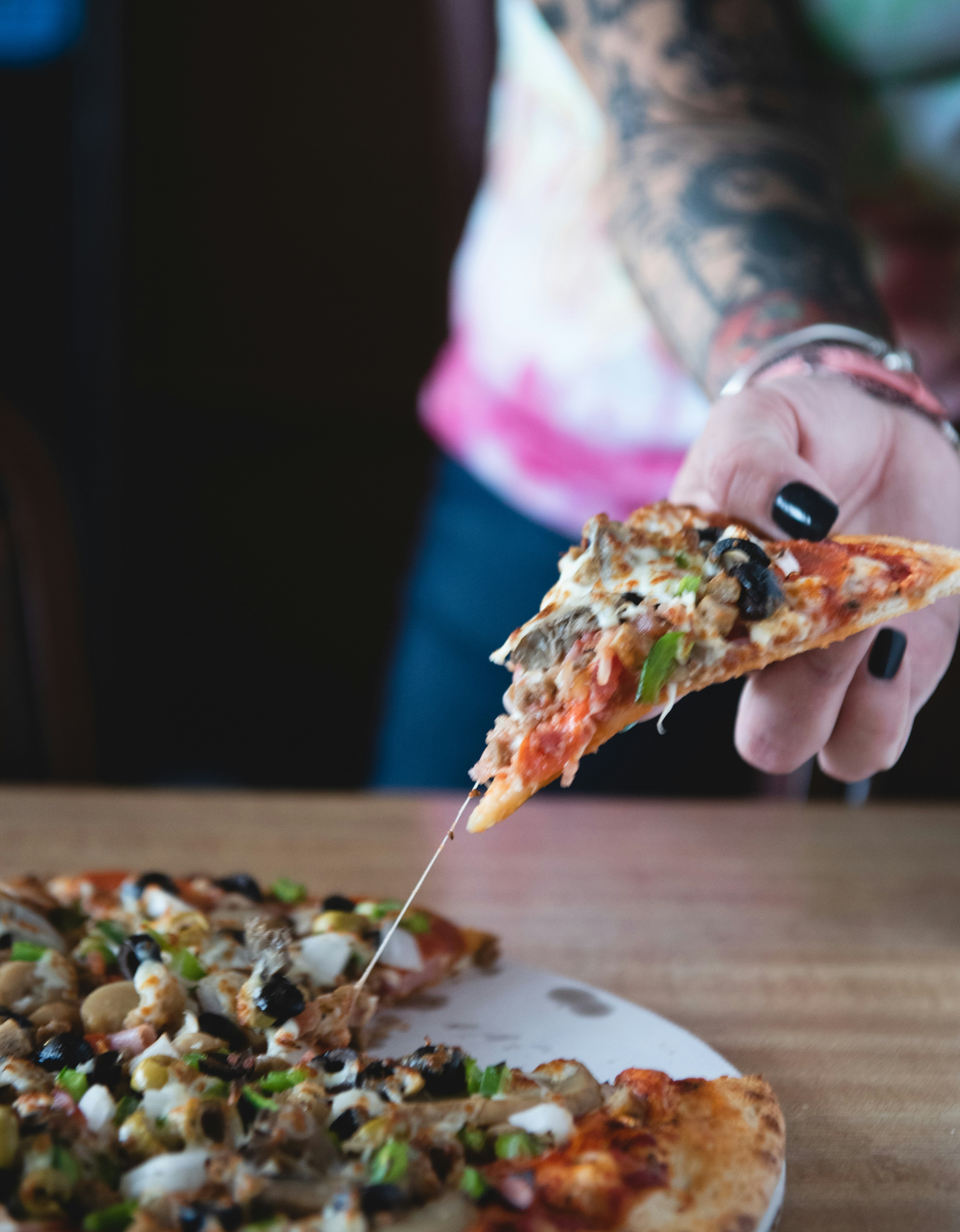 A tattooed person with black nail polish holds a slice of pizza loaded with various toppings like olives and green peppers. The pizza is on a wooden table, and cheese is seen stretching from the slice to the pizza base.