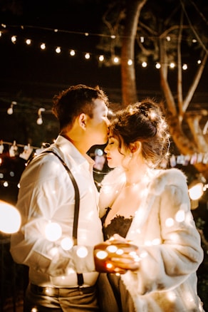 A romantic scene with a couple enveloped in warm, glowing string lights. The man gently kisses the woman's forehead while she holds some lights in her hands. Both are dressed elegantly, and the background is softly lit, creating an intimate atmosphere.