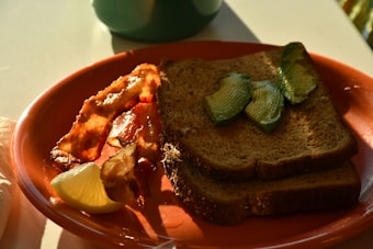 A breakfast plate contains crispy bacon, two slices of whole grain toast with avocado slices, and a wedge of lemon. The plate is a warm terra cotta color, and sunlight casts a natural glow over the food, evoking a cozy and inviting morning ambiance.