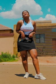 A person stands confidently on a paved outdoor surface, wearing a white tank top with the words 'Fan-Flippen-Tastic' and a black skirt with floral embroidery. Sneakers complete the outfit, and the person is making a playful facial expression. A clear blue sky with fluffy white clouds forms the backdrop, along with a residential building with a roof and a striped wooden gate.
