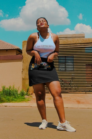 A person stands confidently on a paved outdoor surface, wearing a white tank top with the words 'Fan-Flippen-Tastic' and a black skirt with floral embroidery. Sneakers complete the outfit, and the person is making a playful facial expression. A clear blue sky with fluffy white clouds forms the backdrop, along with a residential building with a roof and a striped wooden gate.