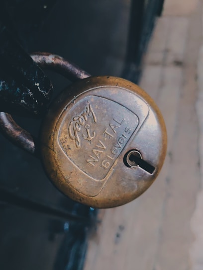 A close-up view of an old, brass padlock with the brand name and specifications engraved on its surface. The lock is attached to a metal latch and has a slightly worn appearance.