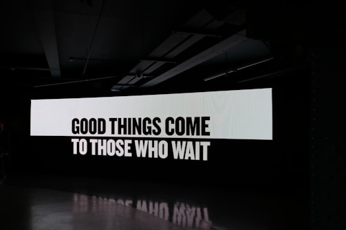 A large digital screen displays the phrase 'Good things come to those who wait' in bold, black letters on a white background. The surrounding environment is dark, emphasizing the illuminated text.