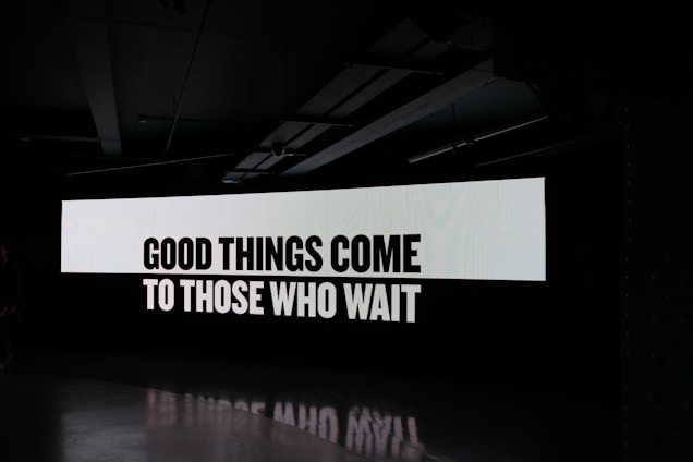A large digital screen displays the phrase 'Good things come to those who wait' in bold, black letters on a white background. The surrounding environment is dark, emphasizing the illuminated text.