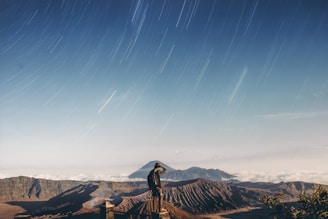 A person stands on a lookout platform, gazing at volcanic mountains under a sky filled with star trails. The mountainous landscape is surrounded by clouds, creating a mystical atmosphere.