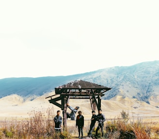 Four people are standing under a wooden structure in a vast open landscape with mountains in the background. The scene includes dry grass and shrubbery in the foreground, with a clear sky overhead.