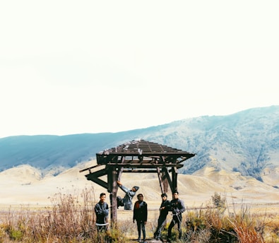 Four people are standing under a wooden structure in a vast open landscape with mountains in the background. The scene includes dry grass and shrubbery in the foreground, with a clear sky overhead.