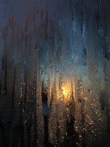 Condensation covers a window, creating droplets and streaks that partially obscure the view outside. The warm glow of sunlight or a lamp is visible through the glass, casting a soft yellow-orange light among the cool tones of the moisture.