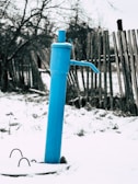 A blue water pump stands prominently against a snowy ground. Behind it, a weathered wooden fence stretches out, with bare trees visible in the background. The atmosphere is cold, suggesting a winter scene.