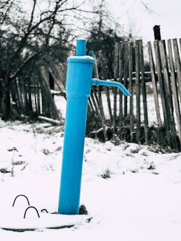 A blue water pump stands prominently against a snowy ground. Behind it, a weathered wooden fence stretches out, with bare trees visible in the background. The atmosphere is cold, suggesting a winter scene.
