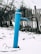A blue water pump stands prominently against a snowy ground. Behind it, a weathered wooden fence stretches out, with bare trees visible in the background. The atmosphere is cold, suggesting a winter scene.