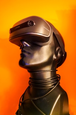 A sleek black mannequin head is adorned with a modern virtual reality headset, against a vibrant orange background. The VR headset is slightly futuristic in design, with smooth curves and a matte finish. A coiled black wire wraps around the neck of the mannequin, adding a sense of technological sophistication.