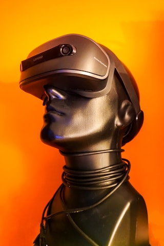 A sleek black mannequin head is adorned with a modern virtual reality headset, against a vibrant orange background. The VR headset is slightly futuristic in design, with smooth curves and a matte finish. A coiled black wire wraps around the neck of the mannequin, adding a sense of technological sophistication.