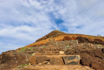 A rugged stone pathway leads up a rocky hillside with a mix of brown, orange, and earthy tones. The vivid blue sky with scattered clouds adds a sense of vastness. Two figures are visible atop the hill, suggesting a hiking or exploration scene.