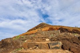 A rugged stone pathway leads up a rocky hillside with a mix of brown, orange, and earthy tones. The vivid blue sky with scattered clouds adds a sense of vastness. Two figures are visible atop the hill, suggesting a hiking or exploration scene.